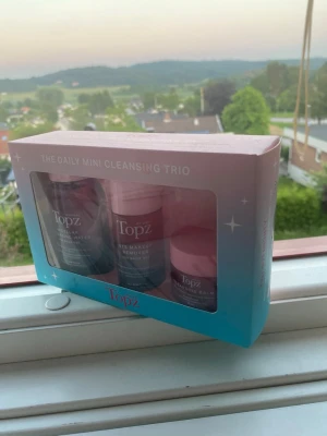 Topz The Daily Mini Cleansing Trio - Säljer ett Topz The Daily Mini Cleansing Trio med tre produkter: micellär rengöringsvatten, ögonmakeup-remover och cleansing balm. Förpackningen är rosa och blå med genomskinligt fönster. Perfekt för smidig hudvård och rengöring.