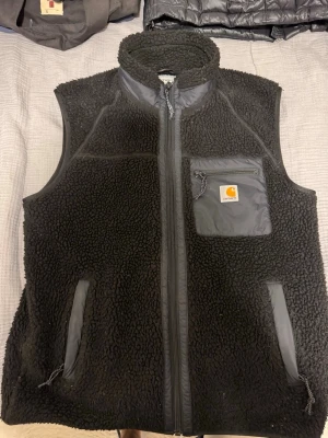 Svart fleeceväst från Carhartt - Svart fleeceväst från Carhartt med hög krage, hel dragkedja och tre fickor med dragkedja. Bröstfickan har Carhartt-logga och detaljer i nylon. Perfekt för lager-på-lager och riktigt skön att bära.