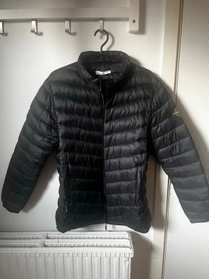 Svart dunjacka med patch på ärmen - Snygg svart dunjacka med quiltad design och hög krage. Jackan har dragkedja framtill och en patch på vänster ärm. Perfekt för kalla vinterdagar och ger en stilren look. Materialet är glansigt och jackan har en lätt puffig form. Säljer pga storlek, jackan är i storlek S men passar även XS