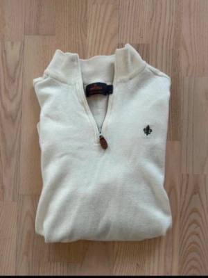 Morris Krämvit tröja med halv dragkedja - Snygg krämvit tröja med halv dragkedja och ståkrage. Tröjan har ett diskret broderat emblem på bröstet och ribbade muddar vid ärmslut och nederkant. Perfekt för lager på lager och enkel att matcha med jeans eller chinos. 