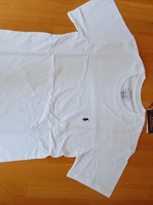 Vit t-shirt från Polo Ralph Lauren - Klassisk vit t-shirt från Polo Ralph Lauren med den ikoniska broderade loggan på bröstet. T-shirten har rund hals och korta ärmar, tillverkad i mjuk bomull för en skön känsla. Perfekt basplagg till garderoben.