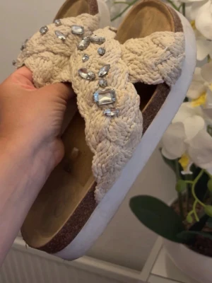 flätade sandaler med strass - Säljer ett par beige sandaler med breda, flätade band i textil och glittrande strassdetaljer. Sulan är vit med en brun innersula. Säljer då dem ej passar mig. 