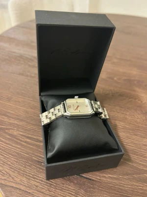 Silverfärgad fyrkantig klocka - Stilren klocka med fyrkantig urtavla och länkarmband i silverfärgad metall. Urtavlan har romerska siffror och tunna visare. Kommer i en svart presentask, perfekt för dig som gillar klassisk och elegant design.