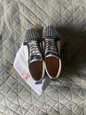 Christian Louboutin sneakers med nitar - Snygga sneakers från Christian Louboutin i svart och vitt läder med blå detaljer. Tåpartiet är täckt av silvriga nitar. Skorna har används men har mycket kvar att ge! Storlek 44 passar dig som vanligtvis har 43. Bara att skriva vid frågor🤝