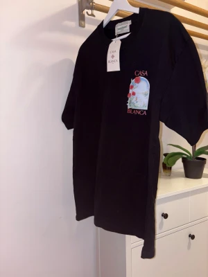 Svart Casa Blanca t-shirt med tryck - Svart t-shirt från Casa Blanca med färgglatt tryck av blommor och landskap både fram och bak. T-shirten har korta ärmar, rund hals och är gjord i mjuk bomull. Perfekt för dig som vill ha en unik och stilren look.
