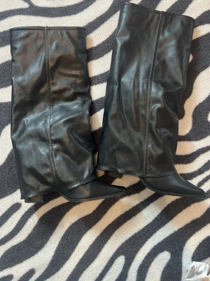 Svarta shark boots från PLT - Säljer ett par svarta shark boots med slouchy passform och spetsig tå. De är tillverkade i skinnimitation och har en låg klack. Stövlarna är knähöga och har en trendig, rynkad look som ger en cool vibe till din outfit. Köpta i vintras från prettylittlething för 850 men aldrig andvända då dem inte riktigt passar, har några små repor fram till på ena skon pågrund av min katt, men syns inte alls när man har på sig dem 