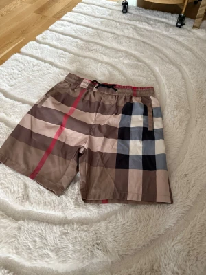 Rutiga badbyxor med beige och rött - Snygga badbyxor med klassiskt rutigt mönster i beige, brunt, svart, vitt och en röd detalj. De har resår i midjan med dragsko och fickor på sidorna samt bak. Perfekta för stranden eller poolen, tillverkade i snabbtorkande material.