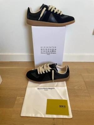 Maison Margiela gats svart - Svarta sneakers från Maison Martin Margiela med beigea skosnören och brun sula. Skorna har en klassisk låg siluett och är tillverkade i en mix av läder och mocka. Perfekta för dig som gillar stilrena och tidlösa sneakers.