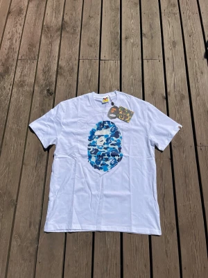 BAPE T-shirt med blå camo Ape Head 👕 -  Helt ny kortärmad BAPE-t-shirt i vitt, med den ikoniska Ape Head-loggan i ett livfullt blått kamouflagemönster på framsidan. Denna klassiska streetwear-tröja levereras med alla originaletiketter kvar, vilket visar att den är i helt oanvänt skick.⭐️ Skick: 10/10 (Helt ny med etiketter) 🎲 Storlek: L 🎨 Färg: Vit med blå camo 📦 Snabb och noggrann frakt