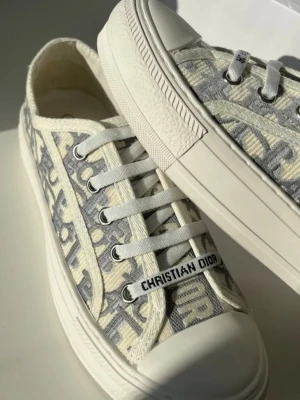 Gråvita Dior sneakers - Snygga sneakers från Christian Dior i grått och vitt med tydligt logomönster över hela skon. Klassisk låg modell med vita snören och robust vit sula. Perfekta för dig som vill sticka ut med en lyxig touch och streetkänsla.