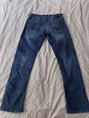 Blå jeans från Replay Grover - Säljer ett par snygga blå jeans från Replay, modell Grover. Jeansen har klassisk femficksdesign, detaljerade sömmar och Replay-logga på fickan. Materialet är slitstarkt denim i en tvättad blå nyans med lätt slitningar för en cool look.