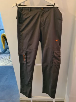 Svarta cargobyxor från Nike - Snygga svarta cargobyxor från Nike med orange logga och detaljer. Byxorna har flera praktiska fickor på benen och är tillverkade i ett lätt, sportigt material. Perfekta för en streetwear-look eller chill vardagsstil.