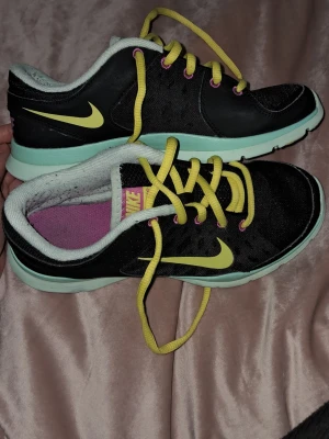 Nike svarta sneakers med gula detaljer - Snygga svarta Nike sneakers med gula skosnören och swoosh-logga. Skorna har en mintgrön sula och lila innersula. Lågt skaft och sportig design i mesh och syntetmaterial. Perfekta för dig som gillar färgglada detaljer och streetstyle. Tvättad