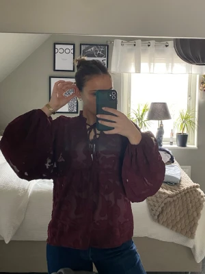 Vinröd blus  - Vinröd blus med strukturmönster och puffiga ärmar från Pernilla Wahlgren. Blusen har knytning vid halsen och lätt tyg som ger en trendig look. 