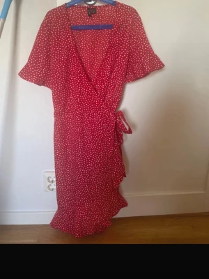 Röd omlottklänning med prickar - Snygg röd omlottklänning med vita prickar och volangkant. Klänningen har korta ärmar, knyt i midjan och v-ringad hals. Perfekt för sommarens alla tillfällen och ger en fräsch och trendig look.