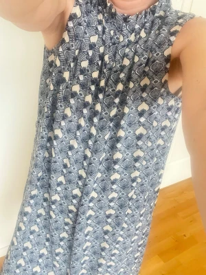 Mönstrad ärmlös klänning i blått och vitt - Säljer en ärmlös klänning med grafiskt mönster i blått och vitt. Klänningen har en lös passform och hög halsringning, perfekt för varma dagar. Materialet känns lätt och luftigt, troligen bomull eller polyester. Supersnygg till sneakers eller sandaler!