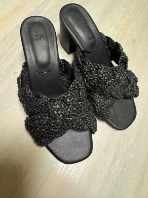 Svarta flätade sandaler med klack H&M - Säljer ett par svarta sandaler från H&M med breda flätade remmar i syntetmaterial och chunky klack. Sandalerna har fyrkantig tå och öppen häl, vilket ger en trendig look. Perfekta för dig som gillar stilrena och moderna skor.