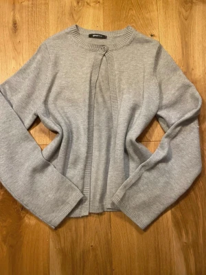Grå kofta med en knapp - Snygg grå kofta från Gina Tricot med rund halsringning och en knapp framtill. Koftan är stickad i mjukt material och har långa ärmar samt ribbade muddar vid ärmslut och nederkant. Perfekt att slänga över en t-shirt eller linne.