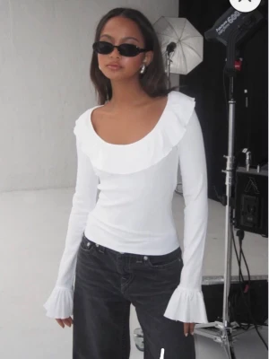 Vit blus med volang och trumpetärm - Supersöt vit blus med bred volang runt ringningen och trumpetärmar. Långärmad och i lätt, vävt material som känns luftigt. Perfekt att styla med jeans eller kjol för en trendig look.