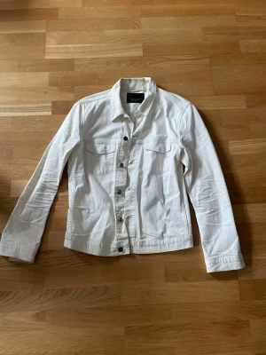 Vit jeansjacka från J.Lindeberg - Snygg vit jeansjacka från J.Lindeberg med klassisk krage, silverfärgade knappar och två bröstfickor med lock. Jackan har raka ärmar och en rak passform som ger en clean och fräsch look. Perfekt att slänga över en t-shirt eller hoodie. Den är solblekt på innekragen.