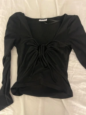 Svart långärmad topp från Noisy May - Trendig svart långärmad topp från Noisy May med snygg snörning framtill och v-ringad hals. Toppen har en croppad passform och är tillverkad i mjukt material som känns skönt mot huden. Perfekt för att lyfta din outfit med en cool detalj.