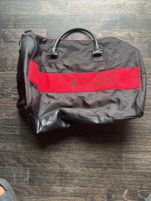 Svart och röd weekendbag Polo Ralph Lauren - Snygg svart weekendbag från Polo Ralph Lauren med bred röd rand och klassisk logga på framsidan. Väskan har dubbla handtag, detaljer i konstläder och rymligt huvudfack. Perfekt för kortare resor eller gymmet.