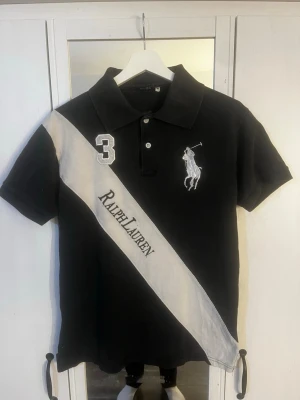 Svart Ralph Lauren t-shirt med tryck - Snygg svart t-shirt från Ralph Lauren med vit diagonal rand och stort tryck av loggan samt siffran 3 på bröstet. Klassisk pikékrage och korta ärmar. Materialet känns som bomull och passformen är normal. Hör av er vid frågor! Lite solblekt.