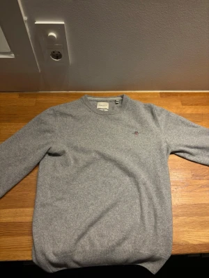 Grå stickad tröja från Gant - Snygg grå stickad tröja från Gant med rund halsringning och diskret broderad logga på bröstet. Tröjan har långa ärmar och ribbade muddar vid ärmslut och nederkant. Perfekt till höst och vinter, och passar till det mesta i garderoben. Material lammull. Storlek M men sitter som S. Litet hål på magen men inget som märks vid användning. 