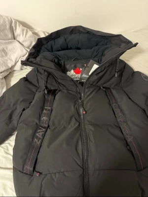 Svart dunjacka från Zavetti Canada - Svart dunjacka från Zavetti Canada med huva och röda detaljer. Jackan har breda band med logotypen på framsidan och en kraftig dragkedja. Perfekt för kalla vinterdagar och har en modern, sportig look.( helt ny)