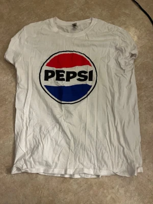 Vit Pepsi t-shirt med tryck - Vit t-shirt med klassiskt Pepsi-logo tryck i rött, blått och svart på bröstet. T-shirten har rund hals och korta ärmar, perfekt för en avslappnad och cool stil.