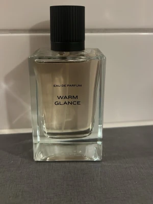 Warm Glance Eau de Parfum - Snygg parfymflaska i glas med minimalistisk design och svart räfflad kork. Flaskan är transparent och visar den ljusa vätskan inuti. Texten 'Eau de Parfum' och 'Warm Glance' står tryckt på framsidan. Perfekt för dig som gillar stilrena dofter.