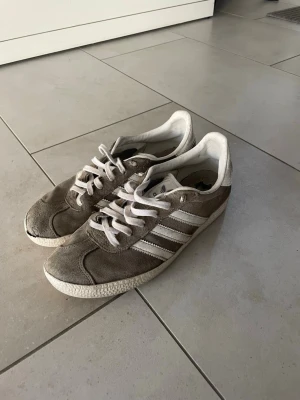 Grå Adidas Gazelle sneakers - Säljer ett par klassiska Adidas Gazelle sneakers i grå mocka med vita detaljer och vita snören. Skorna har den ikoniska tre ränder-designen på sidan och en låg siluett som passar till många olika stilar. Perfekta för dig som gillar retrovibbar och streetwear.
