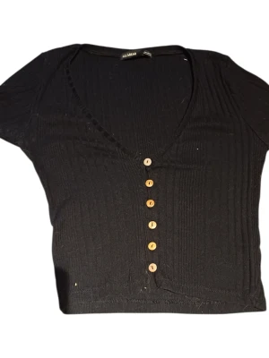 Svart ribbad topp med knappar Pull&Bear - Svart croppad topp från Pull&Bear med ribbad struktur och v-ringning. Toppen har korta ärmar och sex dekorativa knappar framtill. Perfekt till jeans eller kjol för en trendig look.