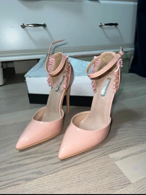 Ljusa rosa pumps med volangdetalj - Säljer ett par ljusrosa pumps från Zara med spetsig tå och hög klack. Skorna har en glansig finish, volangdetaljer längs ankeln och justerbart ankelband med spänne. Perfekta för dig som vill sticka ut med en feminin och trendig look.