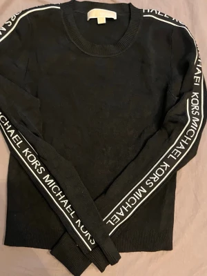 Svart stickad tröja Michael Kors - Snygg svart stickad tröja från Michael Kors med vita logoband längs ärmarna. Tröjan har rund halsringning och ribbade muddar vid ärmslut och nederkant. 