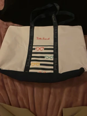 Vit shoppingväska Little Marcel - Stor vit shoppingväska från Little Marcel med mörkblå detaljer och handtag. Väskan har färgglada glasögonmotiv och svarta ränder på framsidan samt broderad logga. Perfekt för att bära allt du behöver till skolan, stranden eller stan.