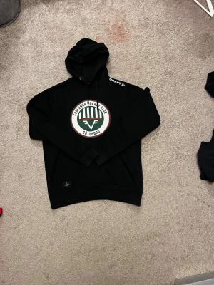 Frölunda HC hoodie - Snygg svart hoodie från Craft med Frölunda Hockey Club Göteborgs logga på bröstet. Klassisk huva med dragsko och tryck på axeln. Tillverkad i mjukt material som passar perfekt för chill eller matchdag. Stora fickor framtill för mobilen eller händerna.