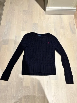 Mörkblå kabelstickad tröja Polo - Kabelstickad mörkblå tröja från Polo Ralph Lauren med rund halsringning och lång ärm. Tröjan har en liten rosa broderad logga på bröstet och bred ribbstickad kant nedtill och vid ärmsluten. Perfekt för en klassisk och stilren look.