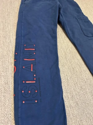 Blå Stone Island mjukisbyxor  - Blå mjukisbyxor från Stone Island med coolt tryck längs ena benet i svart och rött. Byxorna har ribbade muddar vid benslut och en praktisk sidoficka. Perfekta för chill dagar och har en avslappnad passform.