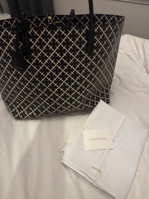 Svart shoppingväska By Malene Birger - Stilren shoppingväska från By Malene Birger i svart med vitt grafiskt mönster. Väskan har två svarta handtag och detaljer i läder. Perfekt för dig som vill ha en rymlig och trendig väska med snygg design. Detta är modellen tote bag. Ny pris 3200kr