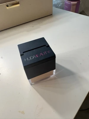 Huda Beauty puder - Puder från Huda Beauty i en fyrkantig transparent burk med svart lock. Kommer med en mjuk applikator för enkel applicering. Färgen är ljusbeige och förpackningen känns lyxig och modern.
