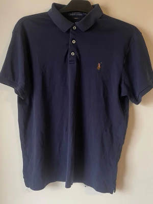 Marinblå piké från Ralph Lauren - Klassisk marinblå pikétröja från Ralph Lauren med korta ärmar och tre knappar framtill. Ikoniska broderade loggan på bröstet. Tillverkad i mjuk bomull som känns skön mot huden. Perfekt till jeans eller shorts för en clean och stilren look. Barn storlek tror jag 👍lite sydd man ser på bilden