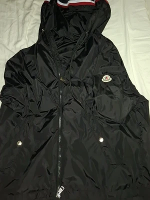 Svart Moncler vindjacka med huva - Snygg svart vindjacka från Moncler med luva och dragkedja framtill. Jackan har två fickor med tryckknappar och Moncler-logga på armen. Detaljer i vitt och rött på huvkanten ger en sportig touch. Tillverkad i glansigt, lätt material som står emot vind.