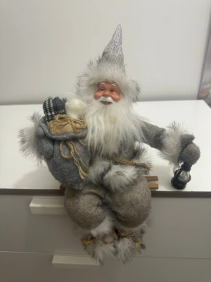 Dekorativ tomte med lykta och säck - En mysig tomtefigur i grå och vita toner, klädd i filt och pälsimitation. Tomten har en spetsig luva, bär en säck med små paket och håller en svart lykta i handen. Perfekt som juldekoration på hyllan eller byrån.