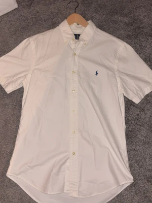 Vit kortärmad skjorta från Polo Ralph Lauren - Vit skjorta från Polo Ralph Lauren med korta ärmar och klassisk button-down krage. Skjortan har broderad blå logotyp på bröstet och knappar framtill. Perfekt för dig som gillar stilrena plagg med ikonisk design.