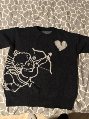 Svart stickad tröja med amor-motiv - Cool svart stickad tröja med vit amor och hjärta på framsidan. Tröjan har rund halsringning och långa ärmar. Perfekt för dig som gillar unika och lekfulla plagg med grafiska tryck.