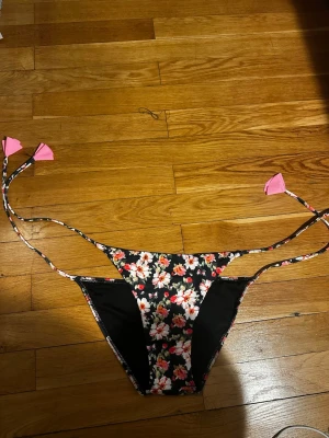Blommiga bikiniunderdelar med knyt - Snygga bikiniunderdelar med svart botten och blommigt mönster i vitt, rosa och rött. Knytband i sidorna med rosa detaljer. Perfekt för stranden eller poolen och ger en trendig look.