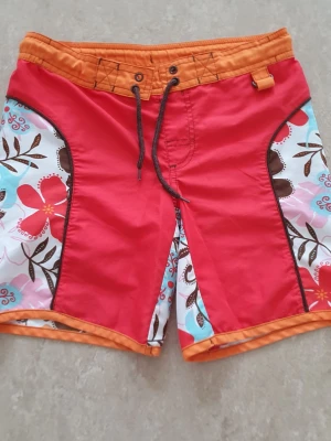 Badshorts - Säljer ett par färgglada badshorts med röd front, orange midja och benkant samt blommigt mönster i rött, rosa, blått och brunt på sidorna. Snörning i midjan och cool surfstil. Perfekta för stranden eller poolen! Finns en fläck.