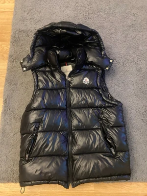 Svart dunväst från Moncler - Snygg svart dunväst från Moncler med glansig finish, avtagbar huva och två dragkedjeförsedda fickor. Västen har Moncler-logga på bröstet och quiltad design som ger en modern look. Perfekt för lager-på-lager-stil under kyliga dagar.