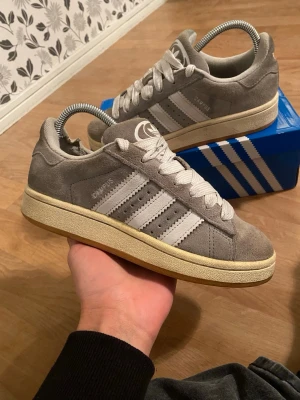 Grå Adidas Campus sneakers i mocka - Snygga Adidas Campus sneakers i grå mocka med klassiska vita ränder på sidorna och vit sula. Skorna har lågt skaft och vita skosnören. Perfekta för dig som gillar retrostil och vill ha något stilrent till vardagslooken.  Har ingen box kvar!